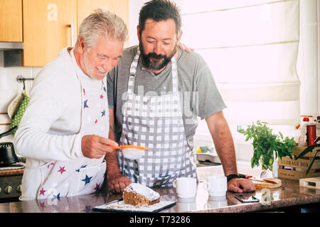 La famiglia a casa a lavorare in cucina con i senior coppia padre e figlio adulto preparare una torta insieme come amici - la diversità e le generazioni miste Foto Stock
