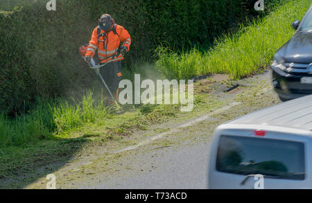 Auto vanno da un lavoratore cittadino la cancellazione della banchina di erba ed erbacce con un mangiatore di erbaccia Foto Stock