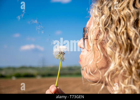 Donna bionda soffia un boom tarassaco e i semi di volare lontano dal cielo blu Foto Stock