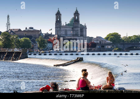 I giovani adulti si trova lungo il fiume Shannon godendo il sole insieme con la Chiesa dei Santi Pietro e Paolo in background Foto Stock