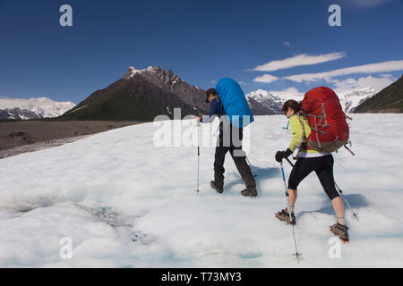 Uomo e donna zaino in spalla attraverso il Root Glacier con ramponi durante l'estate verso Donoho Peak, Wrangell Mountains, Wrangell-St. Elias Nation... Foto Stock