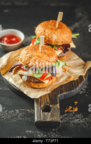 Due deliziosi hamburger fatti in casa a base di carne di manzo, pomodori, formaggi, rucola e bietole su un tagliere di legno. Cucina di strada, fast food. Foto Stock