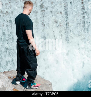 Giovane uomo bello in piedi accanto alla cascata. Blogger e gli ospiti in vacanza. Foto Stock