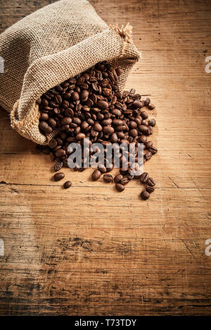 Piena arrosto di miscele di caffè in grani fuoriuscita da un piccolo sacchetto di tela su legno rustico con spazio di copia in formato verticale Foto Stock