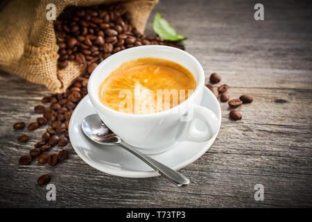 Hot spumoso caffè appena fatto in un bianco tazza e piattino con un sacchetto di fagioli arrosto versarsi sul legno rustico dietro e spazio di copia Foto Stock