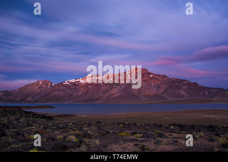 I colori alpenglow la sommità di una catena montuosa in rosso. C'è un lago, rocce vulcaniche e piante del deserto visibile nella parte centrale e di primo piano Foto Stock
