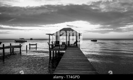 Immagine in bianco e nero di un dock con posti a sedere e ormeggio barche sull'acqua tranquilla al tramonto; Roatan, isole di Bay Reparto, Honduras Foto Stock