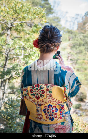 Vista posteriore di una ragazza bellissima da indossare il kimono a scattare foto con il cellulare in un giardino Foto Stock