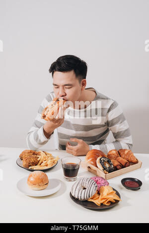 Ritratto di uomo asiatico seduto e snacking ciambelle isolate su sfondo bianco. Uomo grasso concetto di dieta Foto Stock