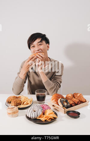 Ritratto di uomo asiatico seduto e snacking ciambelle isolate su sfondo bianco. Uomo grasso concetto di dieta Foto Stock