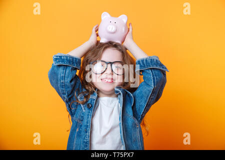 Concetto finanziario di Paghette bambini. Un bambino ragazza con Salvadanaio su un colore di sfondo arancione Foto Stock