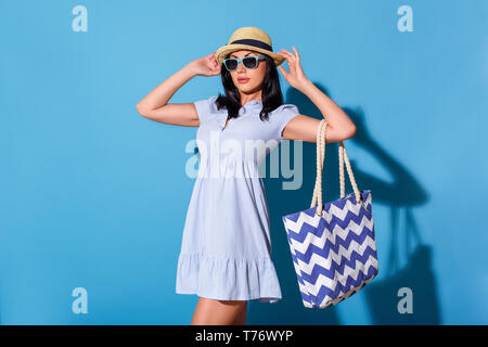Giovane donna abito indossando occhiali da sole e cappello isolato permanente sulla parete blu borsa da trasporto in posa alla telecamera fiducioso Foto Stock