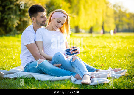 Coppia felice aspetta baby, incinta giovane donna con il marito seduto sul prato verde e azienda Stivali bambino, giovane famiglia e vita nuova nozione Foto Stock