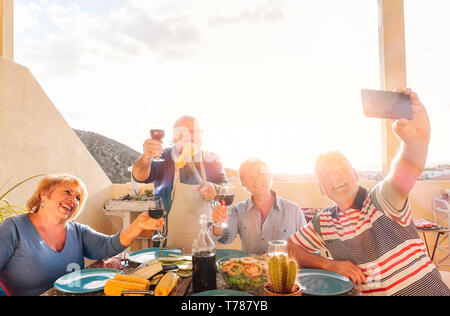 Happy seniors prendendo un selfie con il cellulare e fare un barbecue sul tetto - i pensionati che si diverte a mangiare e a bere vino rosso Foto Stock