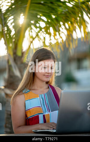 Giovane donna che indossa abiti estivi funziona con computer laptop sotto le palme sulla spiaggia in Florida, Stati Uniti d'America Foto Stock