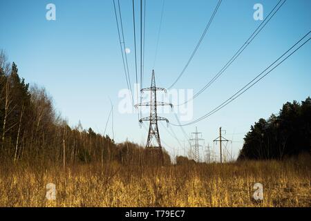 Una linea di alta tensione dei pilastri attraverso un arato campo agricolo, una foresta all'orizzonte ed un cielo blu Foto Stock