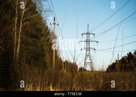 Una linea di alta tensione dei pilastri attraverso un arato campo agricolo, una foresta all'orizzonte ed un cielo blu Foto Stock