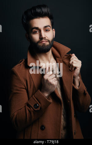 Elegante macho toccando il collare del suo cappotto e posare la fotocamera. Stile, guy ha preparato per autunno.isolato su sfondo nero. Foto Stock