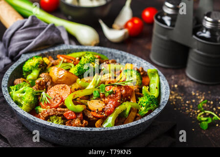 Frittura di carne di manzo stroganoff con patate, broccoli, mais, pepe, carote e salsa in una padella, sfondo scuro. Foto Stock