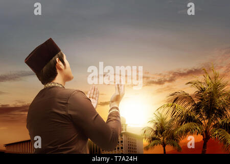 Vista posteriore dei musulmani asiatici man standing e pregando mentre le mani alzate con bella moschea con alberi di palma e il tramonto sul fondo cielo Foto Stock