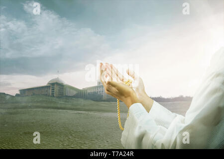 Uomo musulmano di pregare con la preghiera i talloni sulle sue mani sulla duna di sabbia con moschea e scuro dello sfondo del cielo Foto Stock