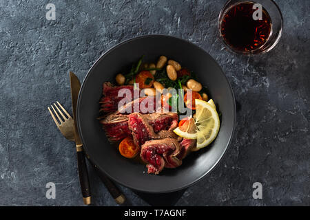 La cena la piastra con carne di manzo con fagioli bianchi, spinaci e pomodori in pietra scura sullo sfondo vista superiore con un bicchiere di vino rosso Foto Stock