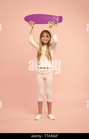 Ragazza con violetta skateboard sopra la sua testa. Campione in street race, infanzia felice concetto. In capretto bianco ponticello, jeans e scarpe da ginnastica isolato su sfondo rosa. Bambino con bella faccia sorridente. Foto Stock