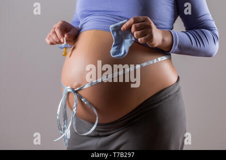 Immagine del ventre della donna incinta con un nastro blu succhietto holding e le calze per bambino su sfondo grigio. Foto Stock