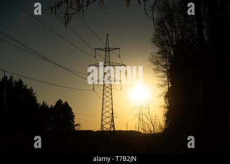 Una linea di alta tensione dei pilastri attraverso un arato campo agricolo, una foresta all'orizzonte ed un cielo blu Foto Stock