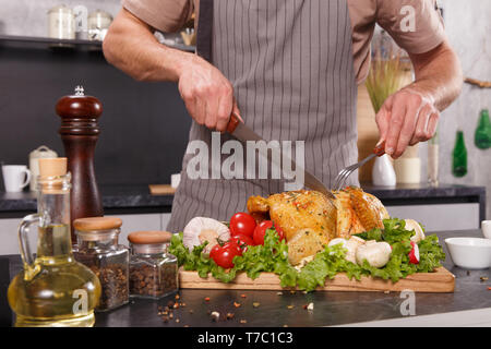 Le mani del capo dell'uomo cuting il pollo al forno con verdure con un grosso coltello su un piatto di legno in un ristorante Foto Stock
