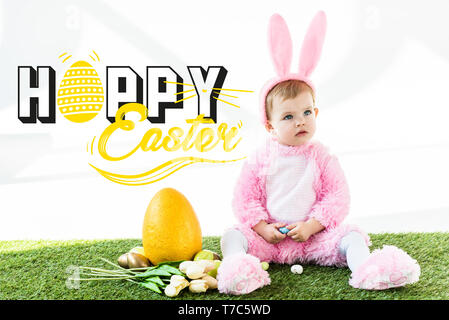 Carino bambino in costume coniglietto seduta vicino colorate uova di pollo, tulipani e giallo uovo di struzzo con felice Pasqua scritte Foto Stock