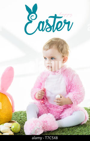 Carino bambino in costume coniglietto seduto sul prato verde con buona Pasqua scritte sopra Foto Stock