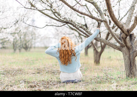 Romantico giovane donna in abito bianco e blu maglione è seduta sul terreno nella primavera del giardino fiorito. Vista posteriore. Concetto di primavera. Foto Stock