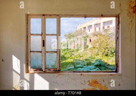 Interno di una casa desolata e un telaio di finestra con il vetro rotto guardando attraverso un prato verde campo Foto Stock