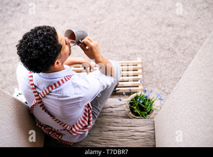 Una vista superiore del giovane uomo grembiule eith seduto davanti alla porta di casa, bere il tè. Foto Stock