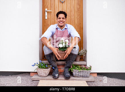 Un Ritratto di giovane allegro uomo seduto davanti alla porta di casa, piantare fiori. Foto Stock