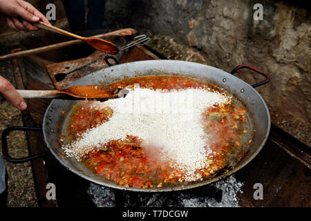 Primo piano di un giovane uomo caucasico preparare una tipica paella spagnola, in una padella per paella con fuoco di legna, all'aperto Foto Stock