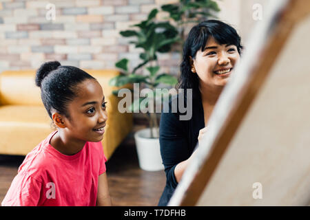 Giovane insegnante dando African American kid una lezione di arte e di insegnare a vernice su cavalletto all'interno del soggiorno in naturale della luce ambientale Foto Stock