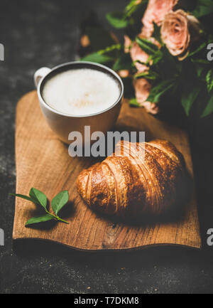 Pausa caffè. Tazza di cappuccino caldo con croissant fresco rustico di legno e bouquet di fiori rosa nero su sfondo squallido. Umore a molla Foto Stock