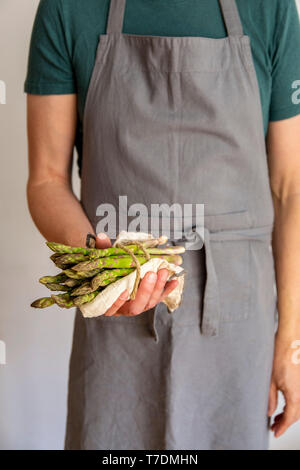 Uomo in grigio grembiule prima di sfondo bianco tenendo un fascio di asparago verde avvolto in un asciugamano nelle sue mani Foto Stock