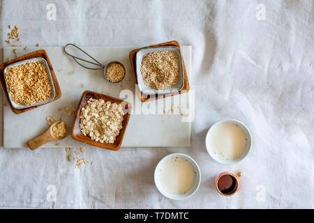 Chicchi d' avena, fiocchi d'avena, oat grist e Latte di avena in ciotole di legno e tazze bianche su marmo bianco bordo su sfondo bianco con spazio di copia Foto Stock