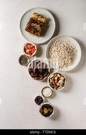 Varietà di frutta secca, noci, miele e fiocchi di avena in ciotole di ceramica per la cottura in casa sana colazione muesli muesli o barrette energetiche su bianco t Foto Stock