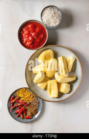 Gli ingredienti per cucinare vegan curry. Raw maturo pelato jackfruit con il bianco riso crudo, tagliate a pezzetti i pomodori e spezie in ceramica bocce su bianco marbl Foto Stock