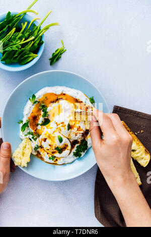 Donna mangiare turco le uova con un po' di pane tostato. Baby spinaci in una piccola ciotola blu accompagna. Foto Stock