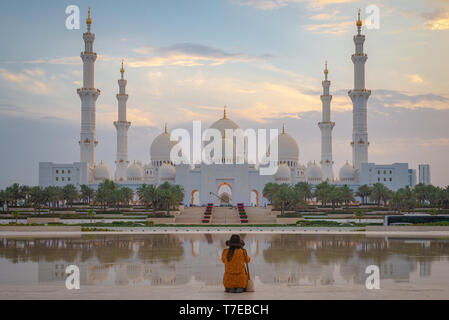 Vista assiale della Grande Moschea di Abu Dhabi al tramonto con la riflessione su uno specchio di acqua con una singola donna che indossa un cappello visto da dietro, Regno un Foto Stock