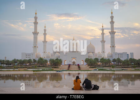 Vista assiale della Grande Moschea di Abu Dhabi al tramonto con la riflessione su uno specchio di acqua con un paio di visto da dietro, Regno emirato Arabo Foto Stock