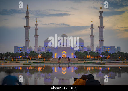 Vista assiale della Grande Moschea di Abu Dhabi al tramonto con la riflessione su uno specchio di acqua con un paio di visto da dietro, Regno emirato Arabo Foto Stock