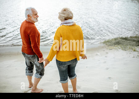 Bella coppia senior vestito in maglioni colorati in piedi insieme sulla spiaggia sabbiosa. Godersi il tempo libero durante il pensionamento vicino al mare Foto Stock