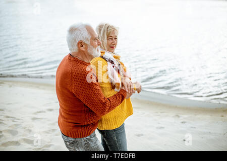 Bella coppia senior vestito in colorate maglioni costeggiata sulla spiaggia sabbiosa, godersi il tempo libero durante il pensionamento vicino al mare Foto Stock