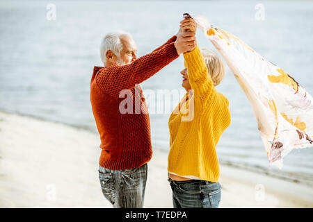 Bella coppia senior giocando con sciarpa durante il tempo ventoso, in piedi insieme sulla spiaggia sabbiosa, godersi il tempo libero durante il pensionamento vicino al mare Foto Stock